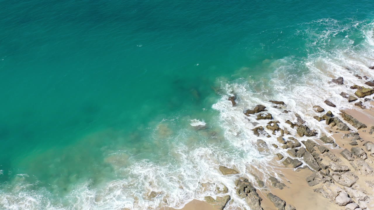 Turquoise water and white surf over rocks beside sand.