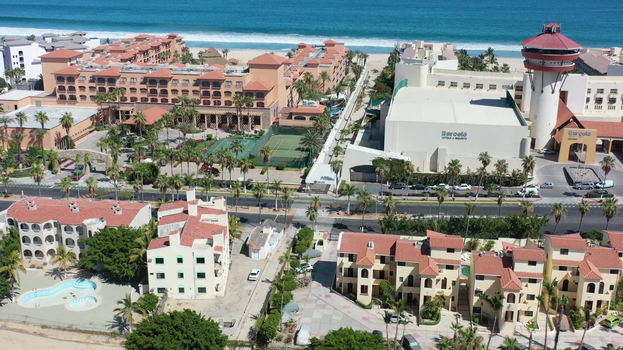 Drone-style aerial frame in Los Cabos, Mexico with a large white hotel building at right, terracotta resort buildings at left, a palm-lined road below, and the ocean beyond.