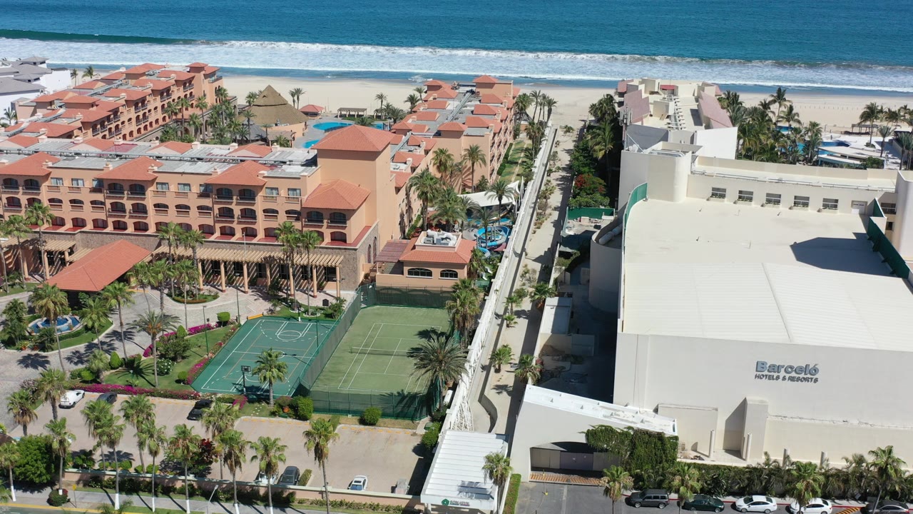 Elevated view in Los Cabos, Mexico showing red-roof hotel buildings, green sport courts, a large white building, sandy beach, surf, and no visible people.