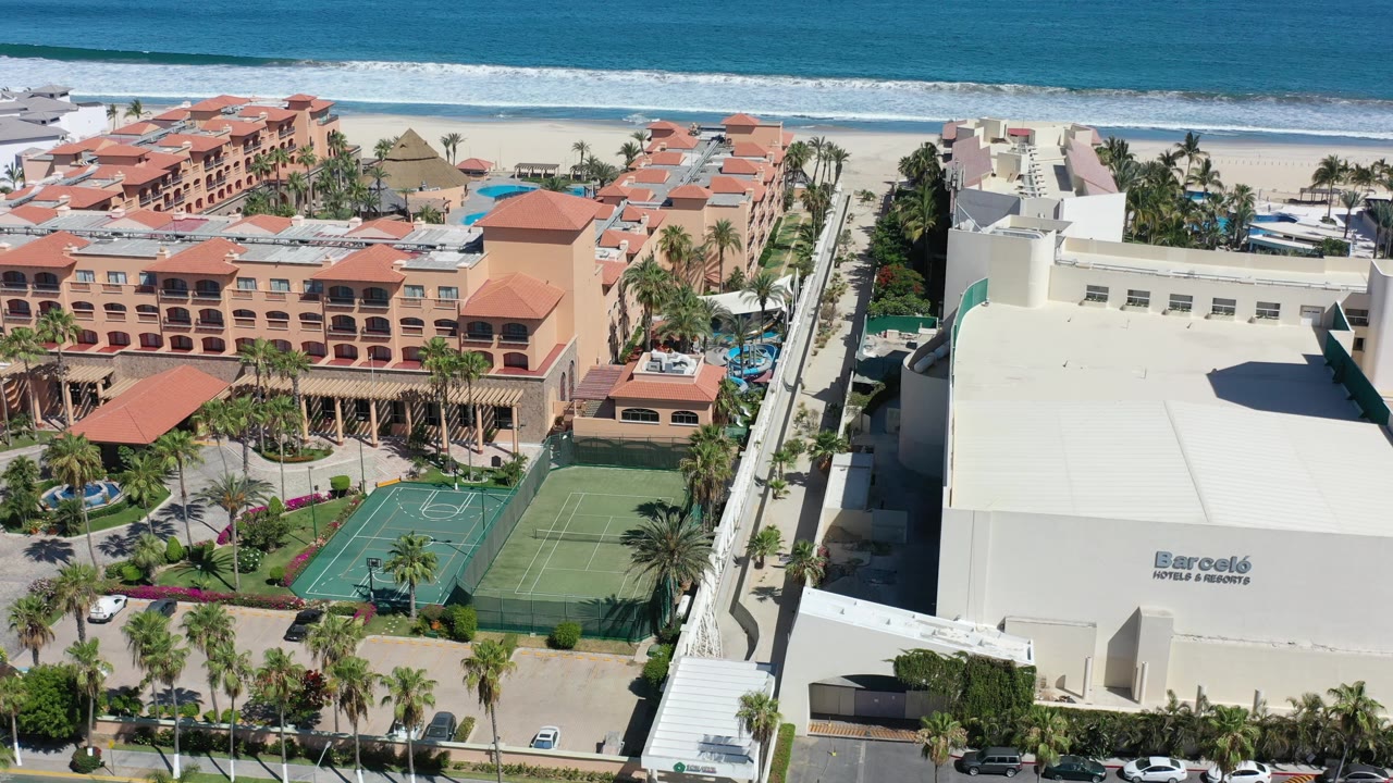 Drone-height view in Los Cabos, Mexico with the red-roof hotel complex centered, sport courts below, beach and ocean at the top, and no visible people.