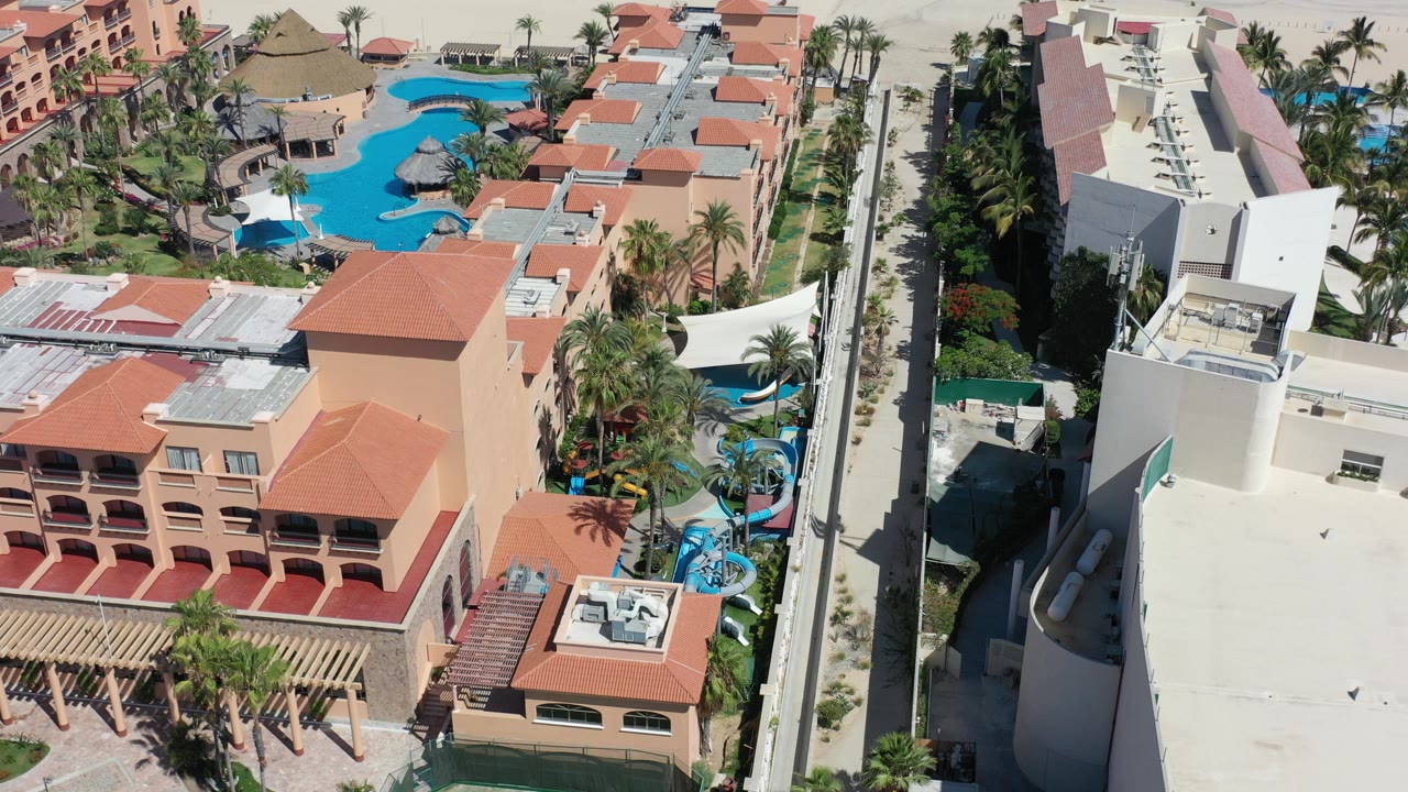 Aerial frame from Los Cabos showing terracotta-roofed villa buildings, bright blue pools, palm landscaping, and a straight road at the right edge.
