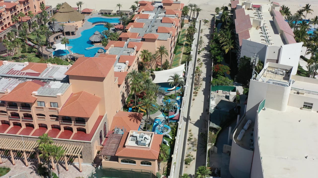 Overhead Los Cabos view with a large curved turquoise pool, orange roof sections, palms, and pale roof surfaces in hard sunlight.