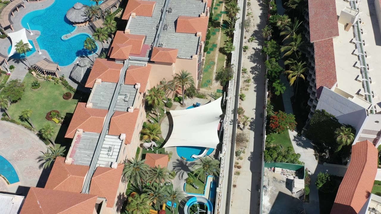 Overhead Los Cabos frame with cream buildings, terracotta roof sections, bright blue pools, palms, walkways, and no visible people.