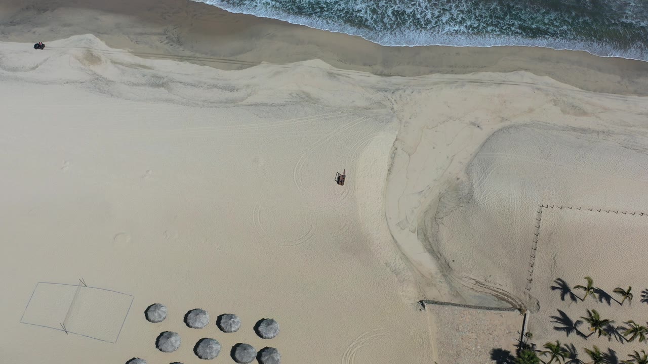 Top-down aerial frame of a Los Cabos beach with white surf at the top, broad pale sand below, small umbrellas near the lower edge, and tiny distant figures or objects near the shoreline.