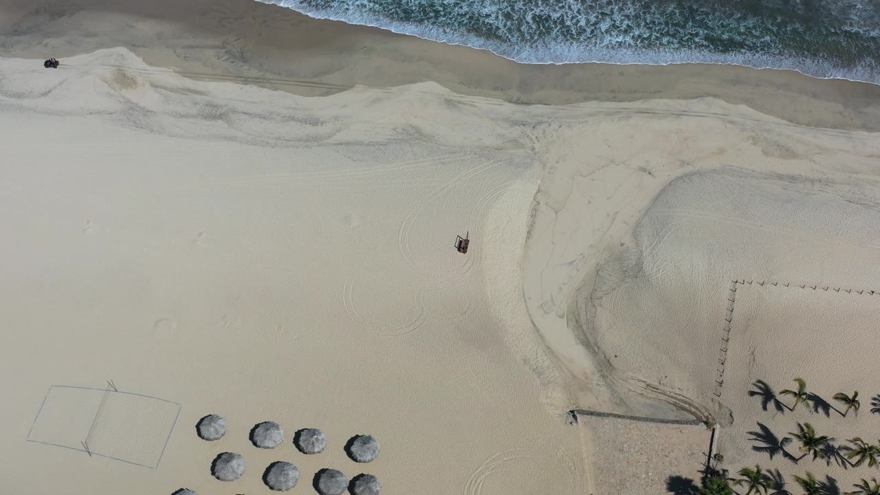 High overhead view in Los Cabos with the surf band across the top, empty-looking sand through the center, and a cluster of umbrellas and palm shadows near the bottom of the frame.