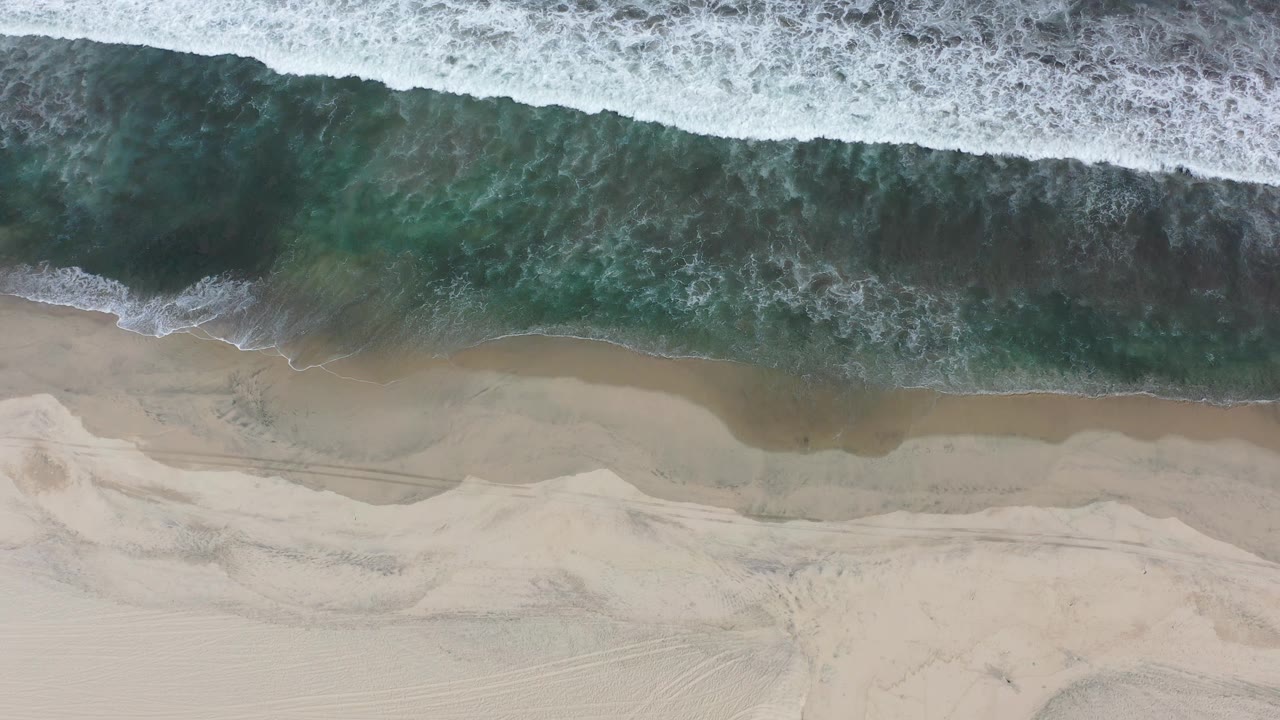 A broad white wave line crosses green-blue water toward a narrow strip of wet sand on the Los Cabos beach.