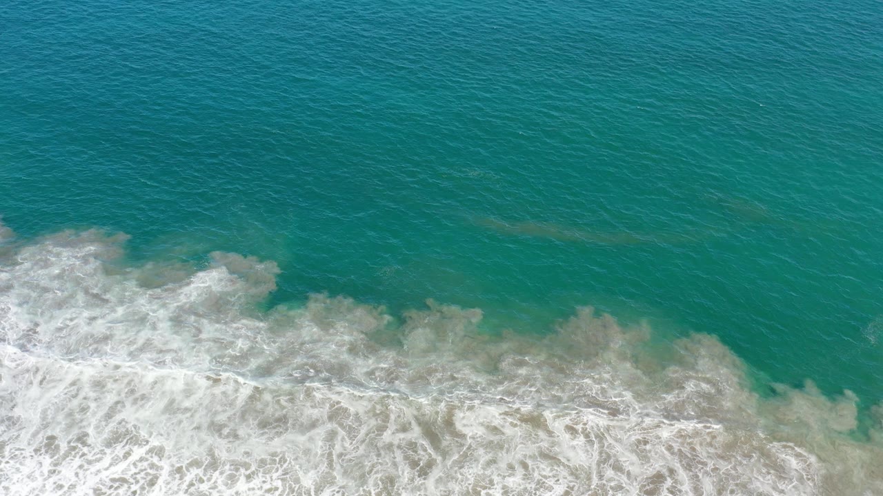 Los Cabos surf seen from above as whitewater spreads across the lower half of the frame under turquoise ocean, no visible people.