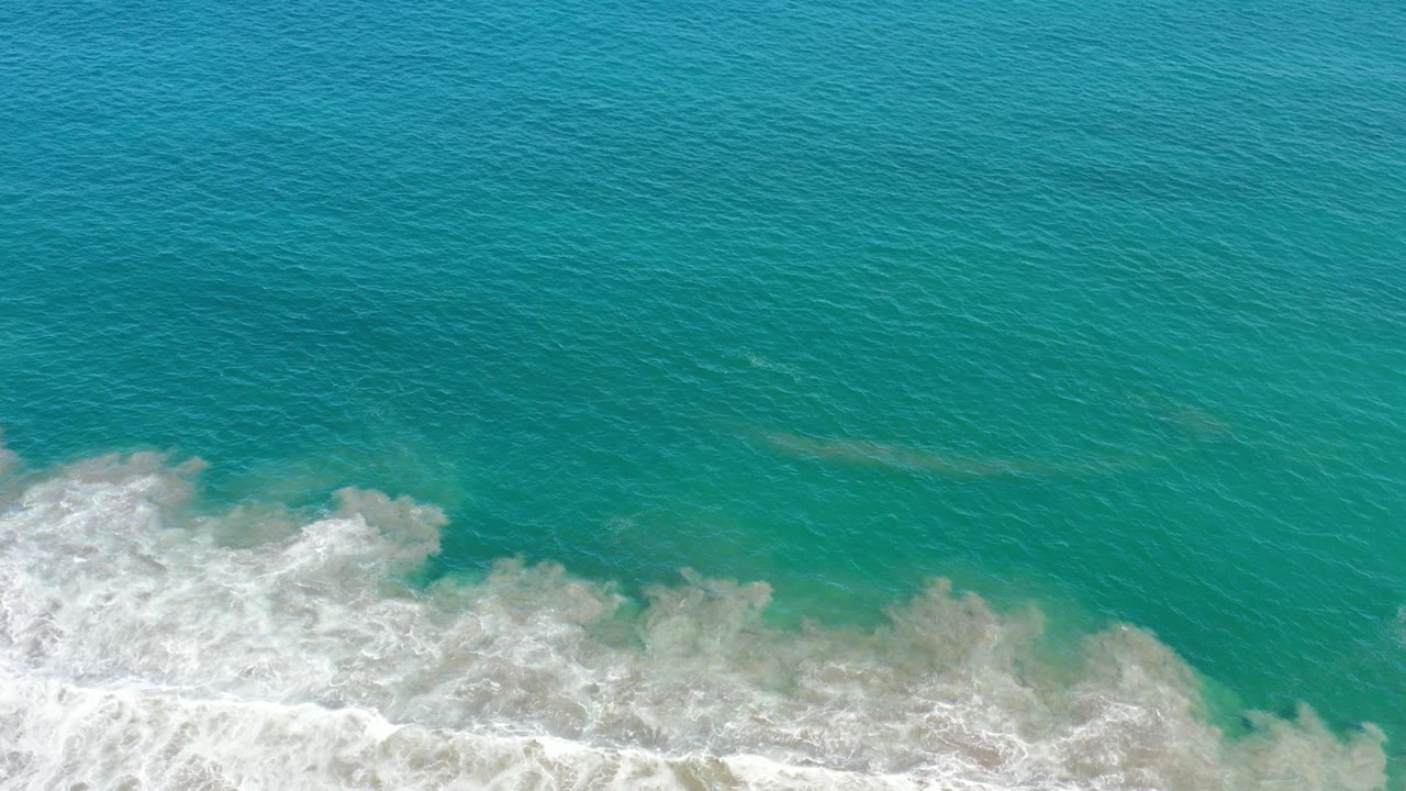 White surf rolls beneath a broad expanse of teal ocean water.