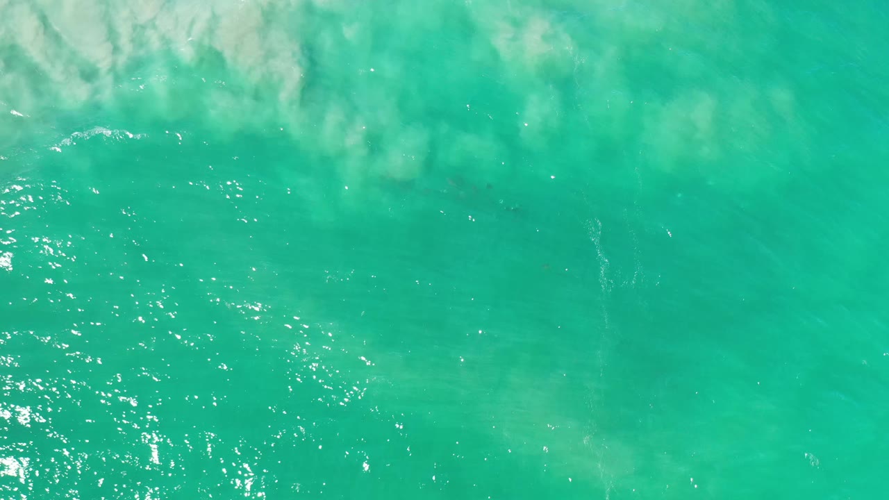 Overhead Los Cabos water view with green-blue color, bright specks of reflected sunlight along the left side, faint cloud reflection above, and no visible people.