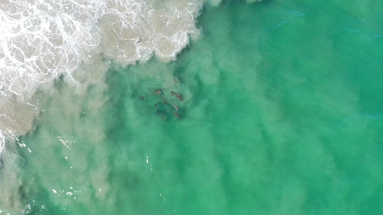 Overhead frame from Los Cabos, Mexico showing white surf entering from the upper left over pale sand, turquoise water filling the right side, and no visible people.