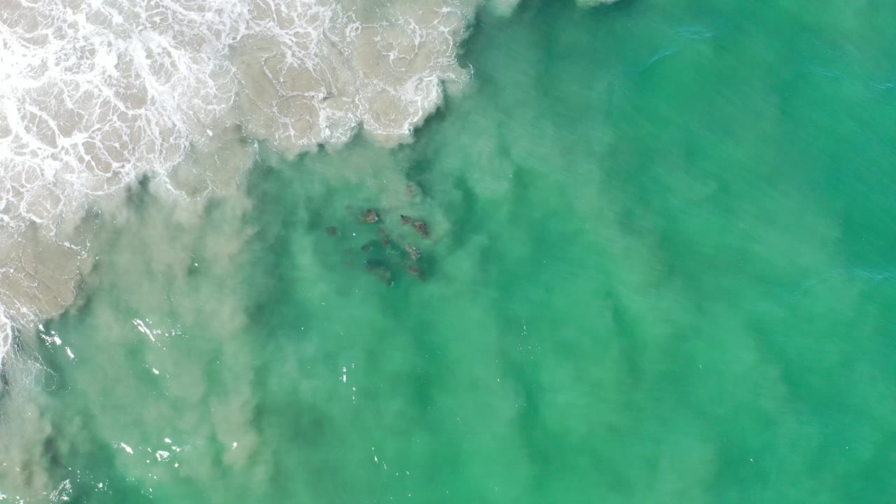 Overhead Los Cabos shoreline view with the foam line slightly farther across the upper-left sand and small dark submerged patches near the center, with no visible people.