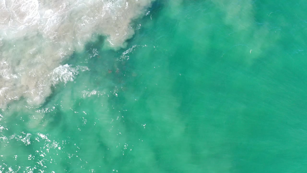 Overhead view of turquoise Los Cabos ocean water with white surf spreading from the upper left corner and no visible people.