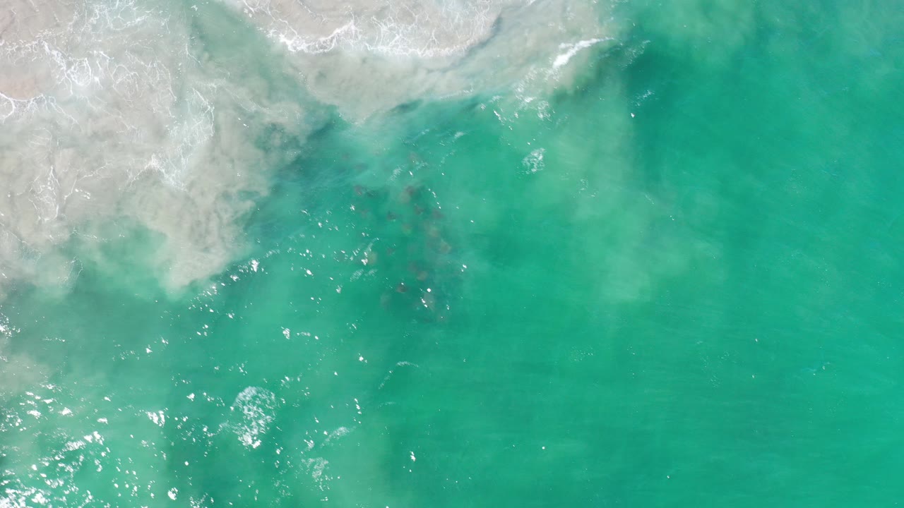 Overhead view of green-blue sea in Los Cabos, Mexico, with white surf and pale sand along the upper edge and no visible people.