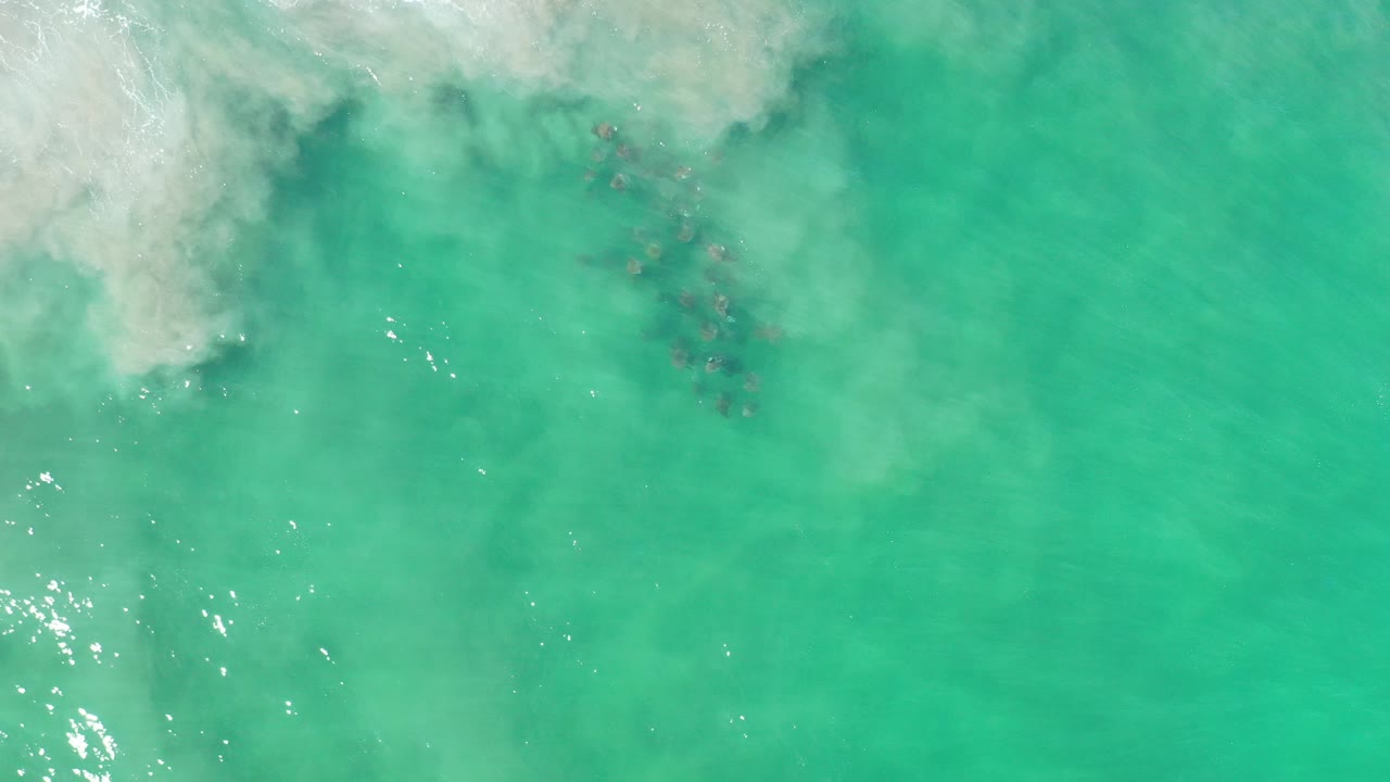 Overhead view of turquoise ocean water in Los Cabos, Mexico, with pale surf foam along the upper left edge, darker submerged patches near center, and no visible people.