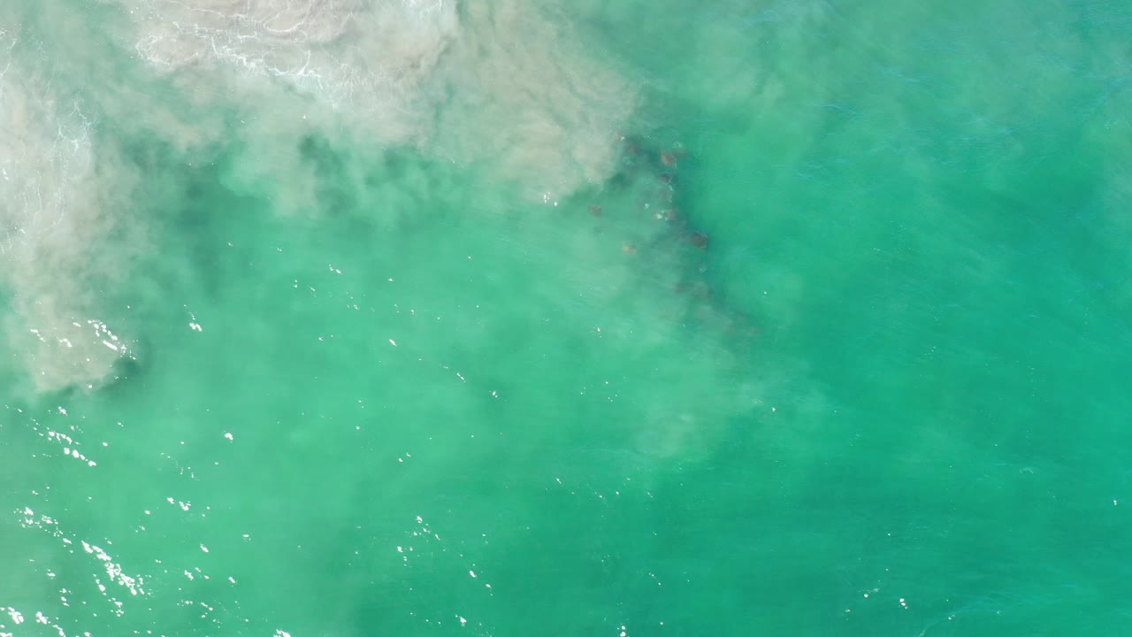Overhead Los Cabos water view with turquoise-green sea, white surf foam pushing in from the upper left, darker patches below the surface, and no visible people.