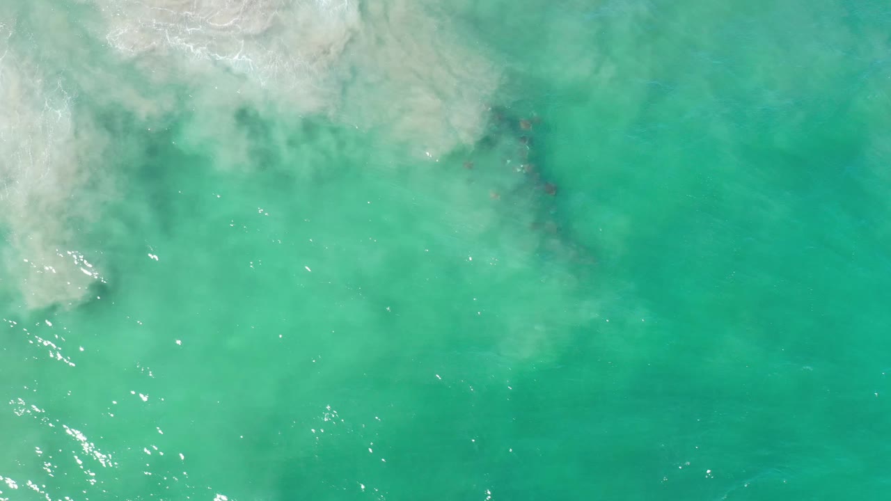 Overhead Los Cabos sea surface with pale foam along the upper left edge, scattered bright reflections near the lower left, and no visible people.