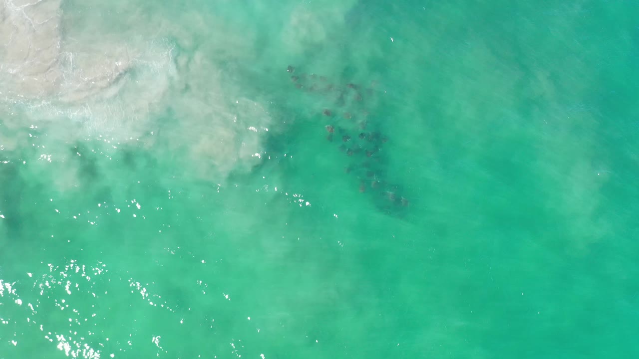 Aerial Los Cabos frame with the pale shallows holding at the upper left, sun reflections near the lower left, darker fish shapes under the center water, and no visible people.