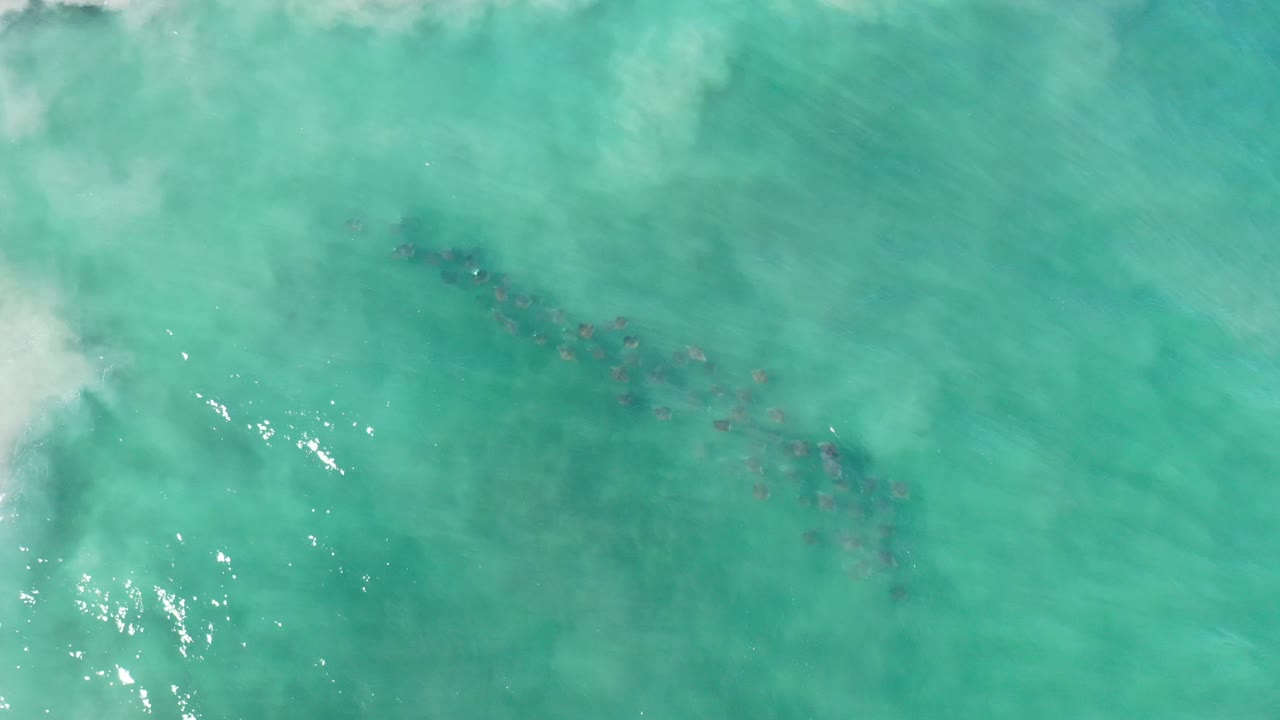 Aerial view over green turquoise water in Los Cabos, Mexico, showing a diagonal dark underwater line, small sun sparkles, and no visible people.