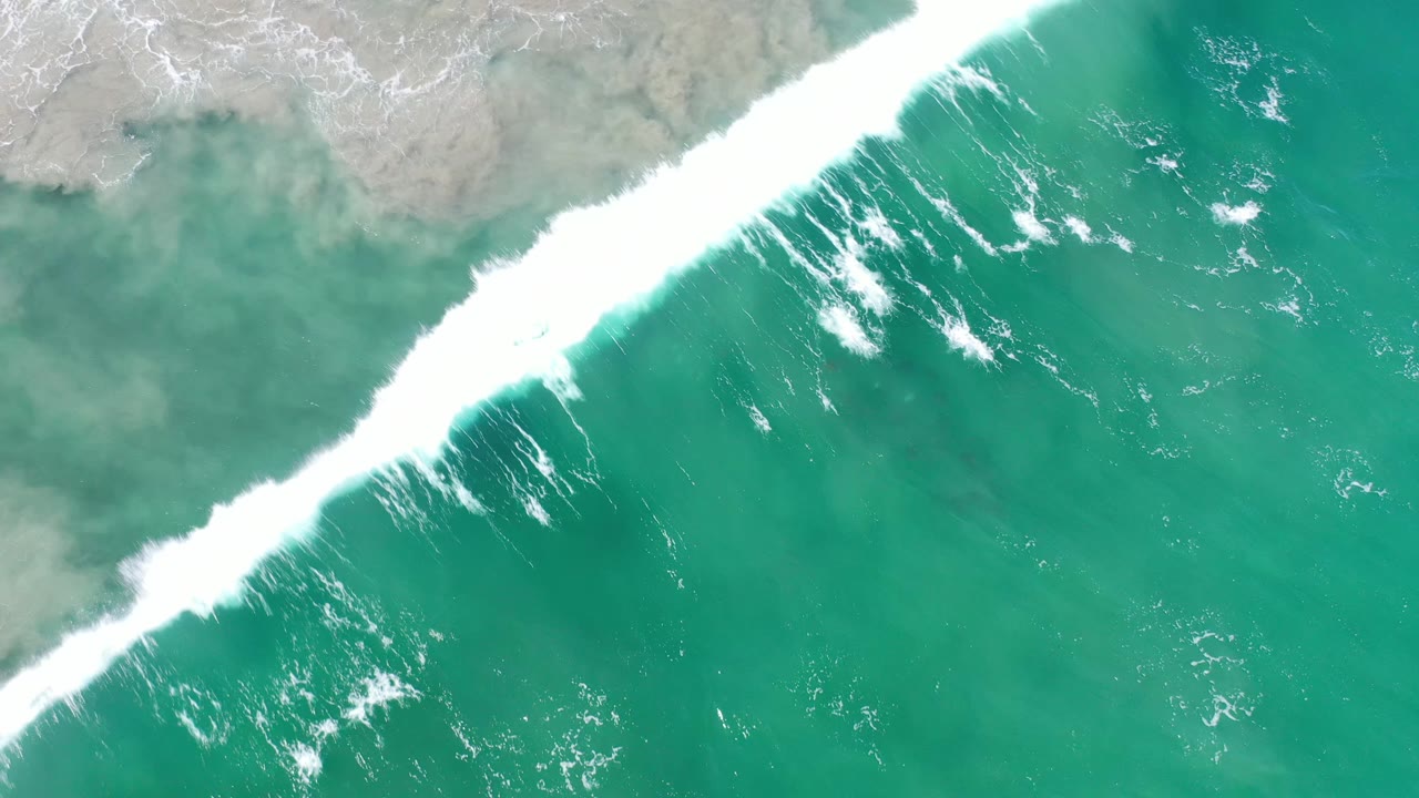 Aerial Los Cabos frame with a bright diagonal wave crest, pale sand under shallow water, and open turquoise sea below, with no visible people.