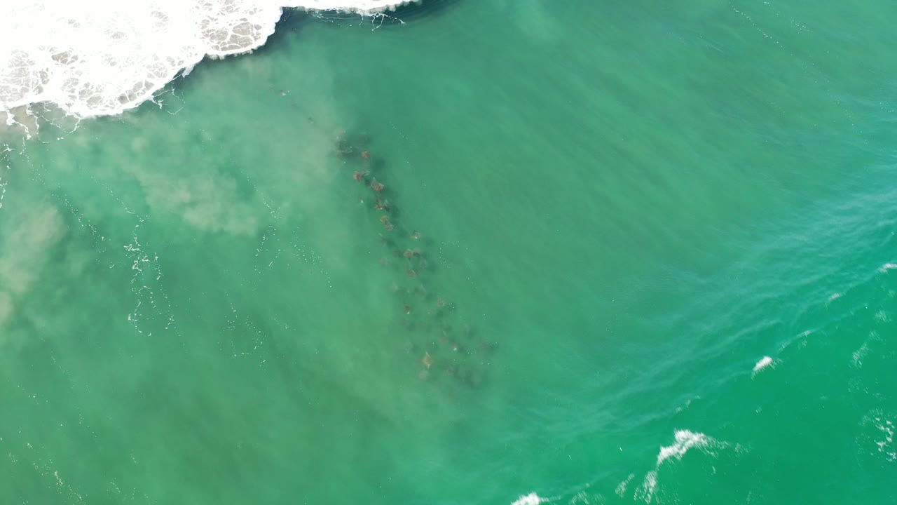 Overhead view of green seawater at Los Cabos with white foam along the upper edge, darker submerged streaks beneath the surface, and no visible people.