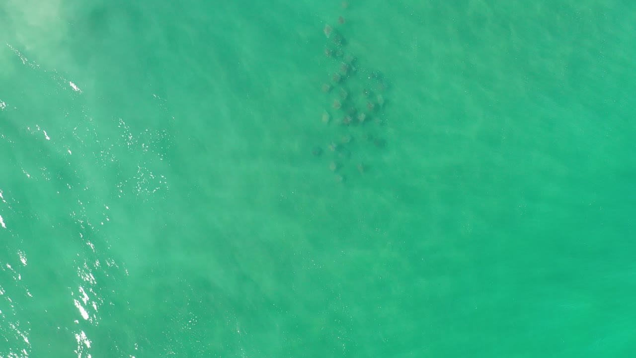 Overhead view of turquoise ocean in Los Cabos, Mexico, with faint darker underwater patches, small ripples, and no visible people.