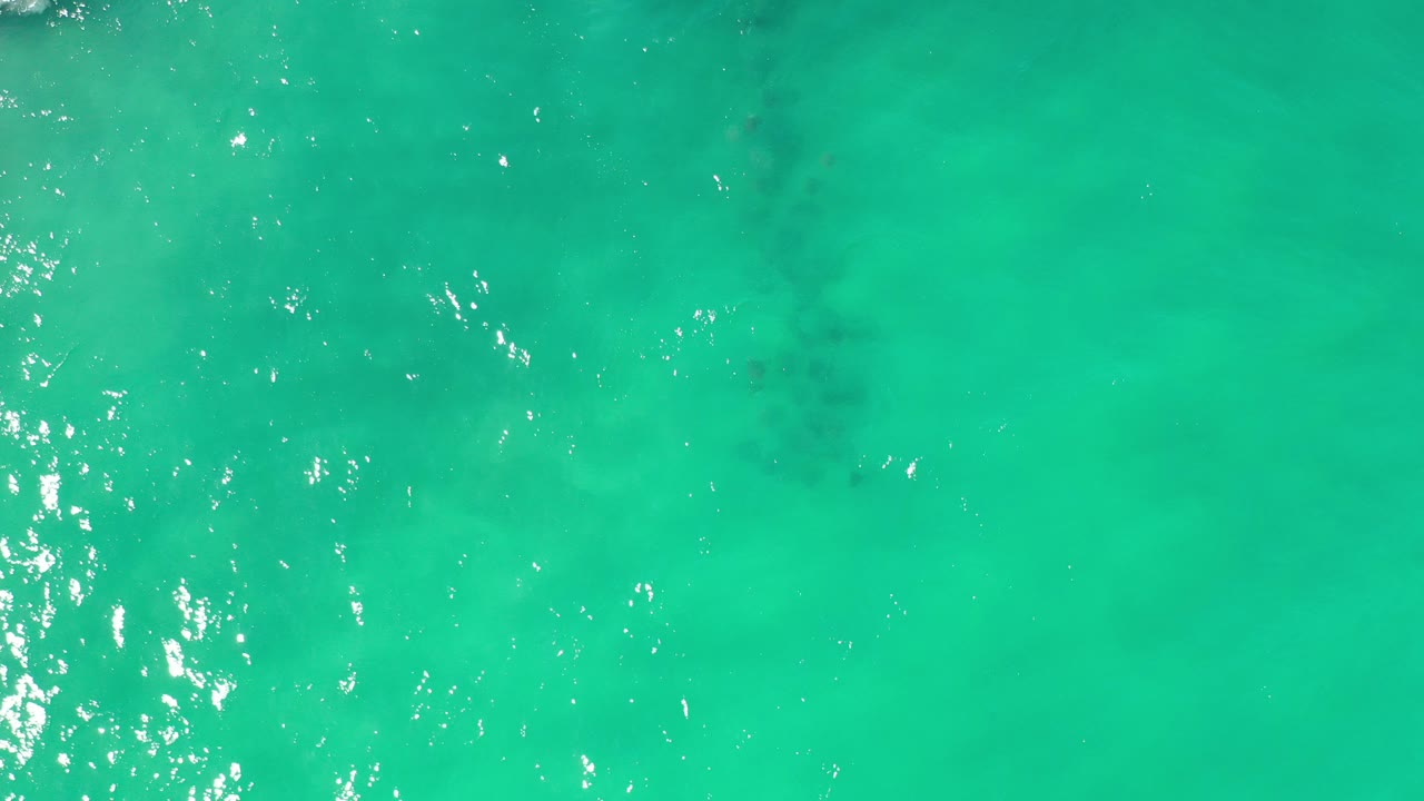 Overhead view of light green water in Los Cabos, Mexico, with bright sun flecks near the left edge, darker submerged patches, and no visible people.
