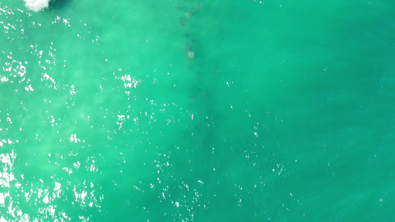 Aerial view over Los Cabos ocean water with a white foam patch at the upper edge and the whale's dark body partly visible beneath the green surface, no visible people.