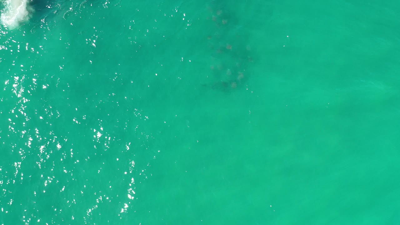 Overhead view of turquoise sea in Los Cabos, Mexico, with bright sun sparkle, faint darker submerged patches, and no visible people.