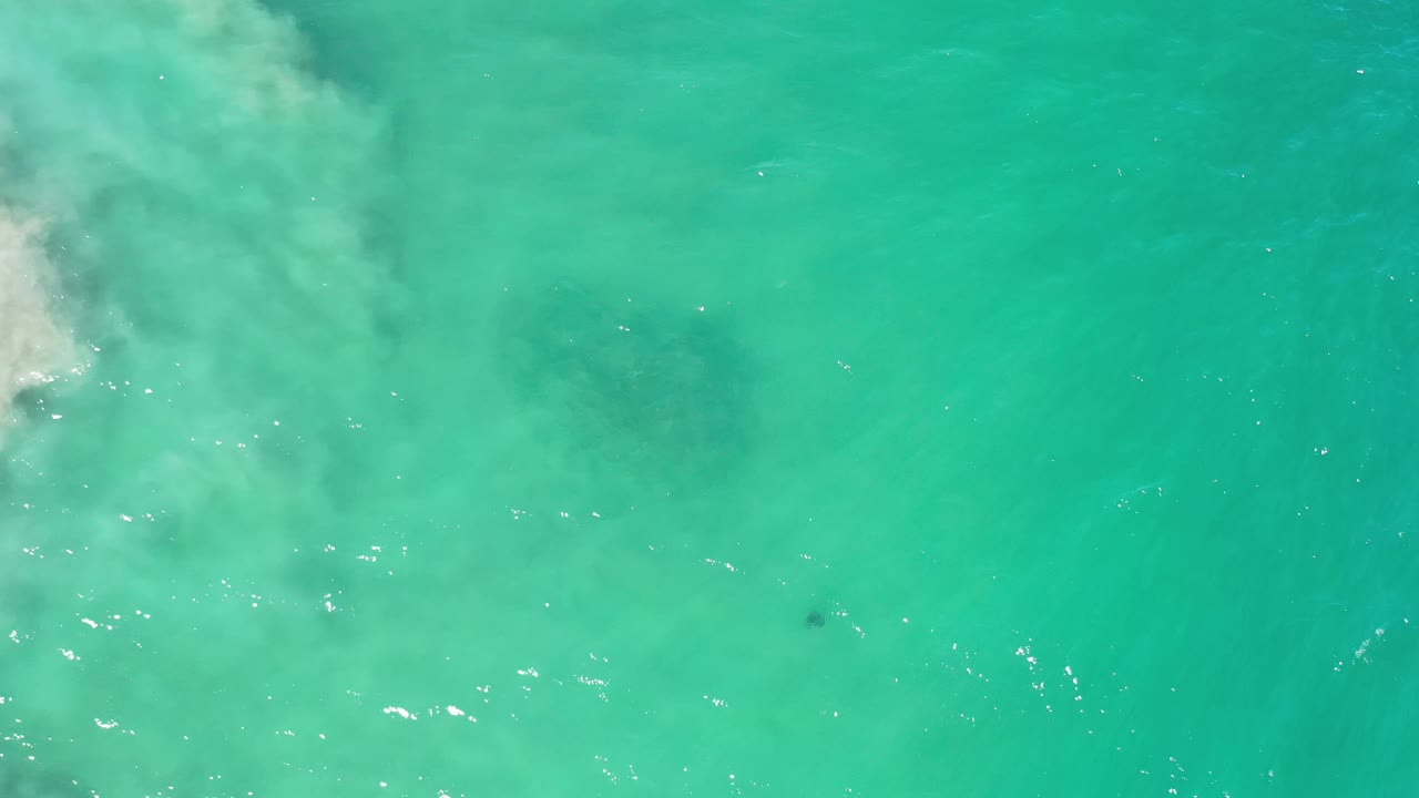 Overhead Los Cabos ocean frame with a larger pale foam patch along the left side, faint sandy haze below the surface, and no visible people.