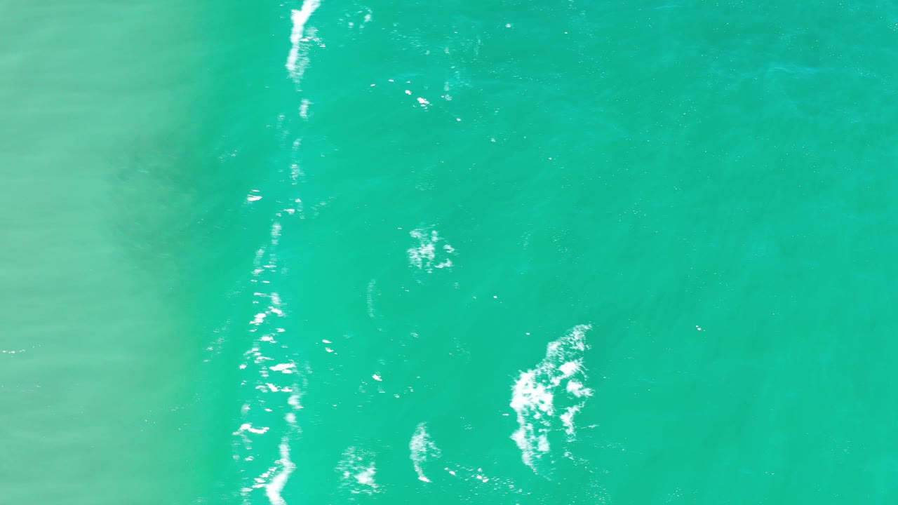 Overhead view of teal ocean water in Los Cabos, Mexico, with a white wave streak along the left side and no visible people.