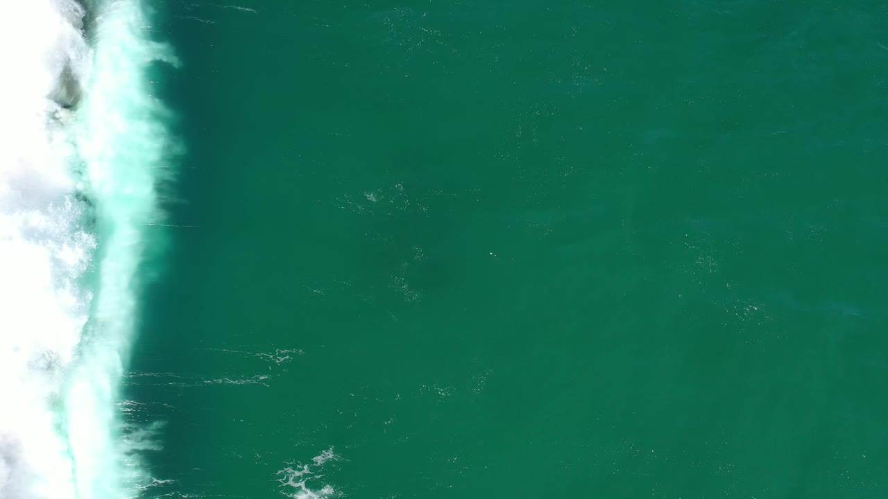 Overhead Los Cabos ocean frame with white wave foam pressing farther down the left margin and open green-blue water to the right, with no visible people.