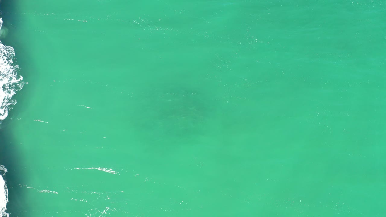 Top-down turquoise ocean with surf along the left edge.