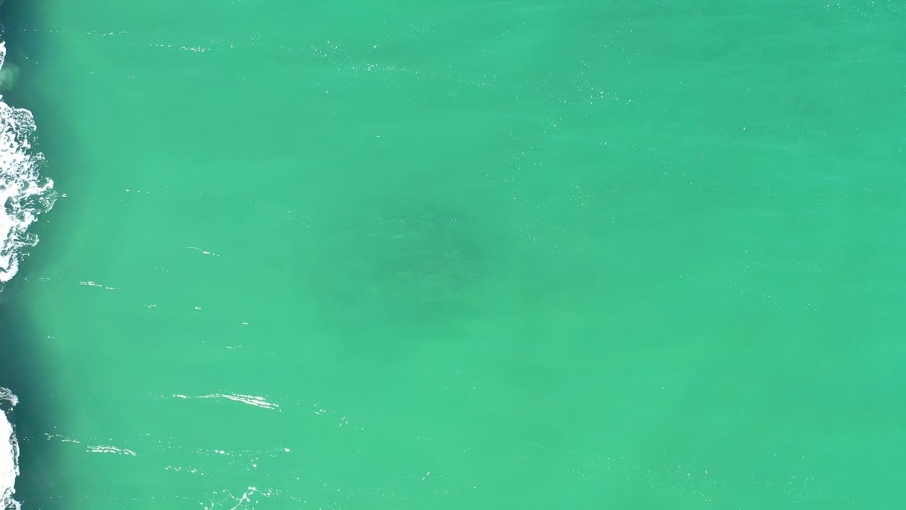 Aerial green ocean water in Los Cabos, Mexico, with faint darker shapes below the surface, white foam along the left edge, and no visible people.