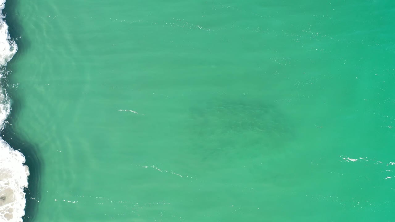Top-down Los Cabos water view showing a thin band of breaking foam on the left, muted green water across the center, faint sediment streaks, and no visible people.