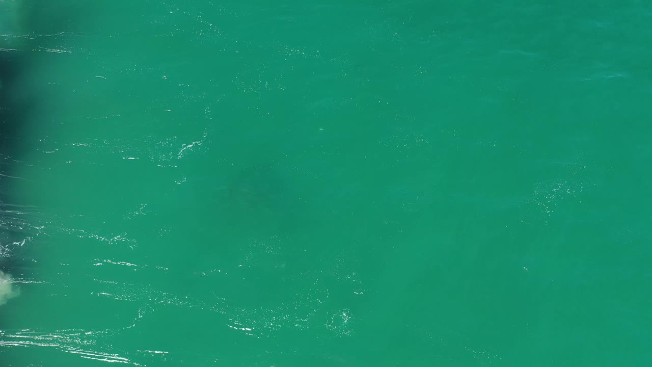 Aerial view of clear green-blue Los Cabos water with thin whitewater lines crossing the surface and no visible people.