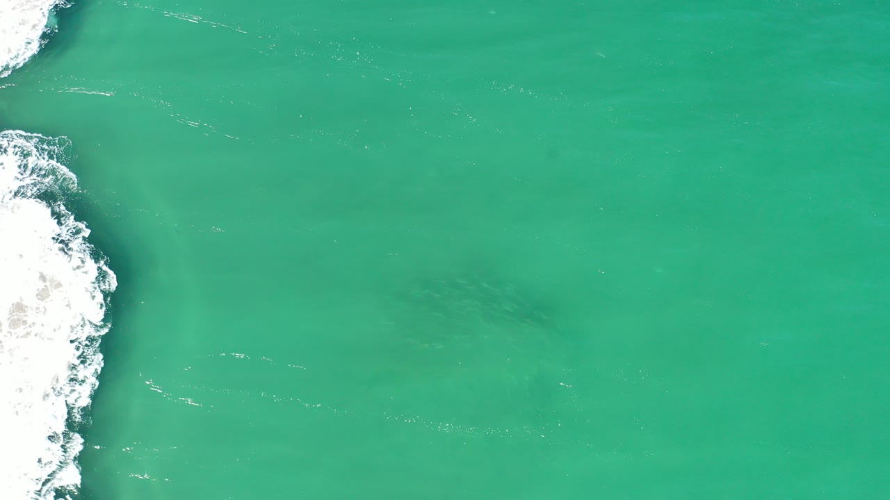 Overhead view of turquoise water in Los Cabos, Mexico, with white surf along the left edge and a faint dark underwater shape near the center, no visible people.