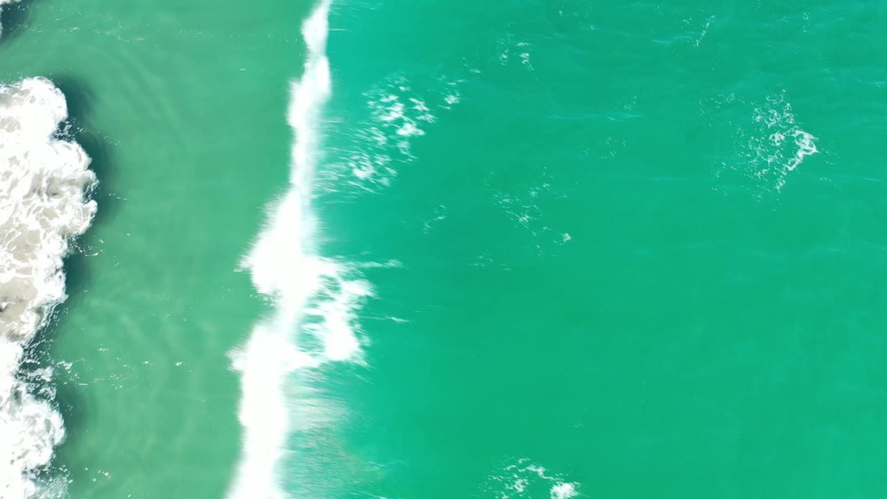 Overhead Los Cabos frame with white foam running down the left-center of the turquoise ocean and rocky shoreline partly visible, with no visible people.