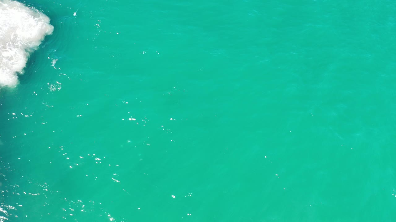 Overhead view of turquoise-green ocean in Los Cabos, Mexico, with white surf at the upper and lower left edges and no visible people.