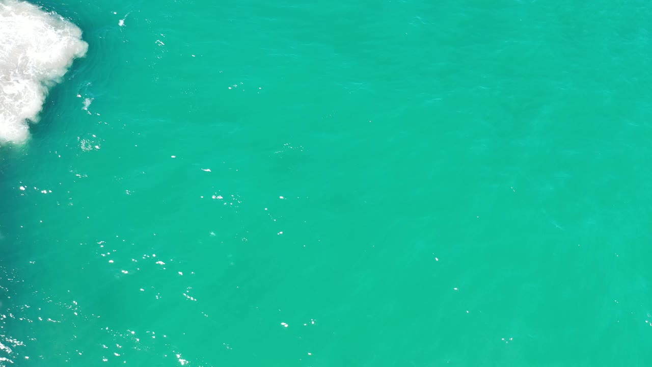 Overhead Los Cabos ocean view with green-blue water, small sunlight reflections near center left, and foamy surf pushing in from the left edge, with no visible people.