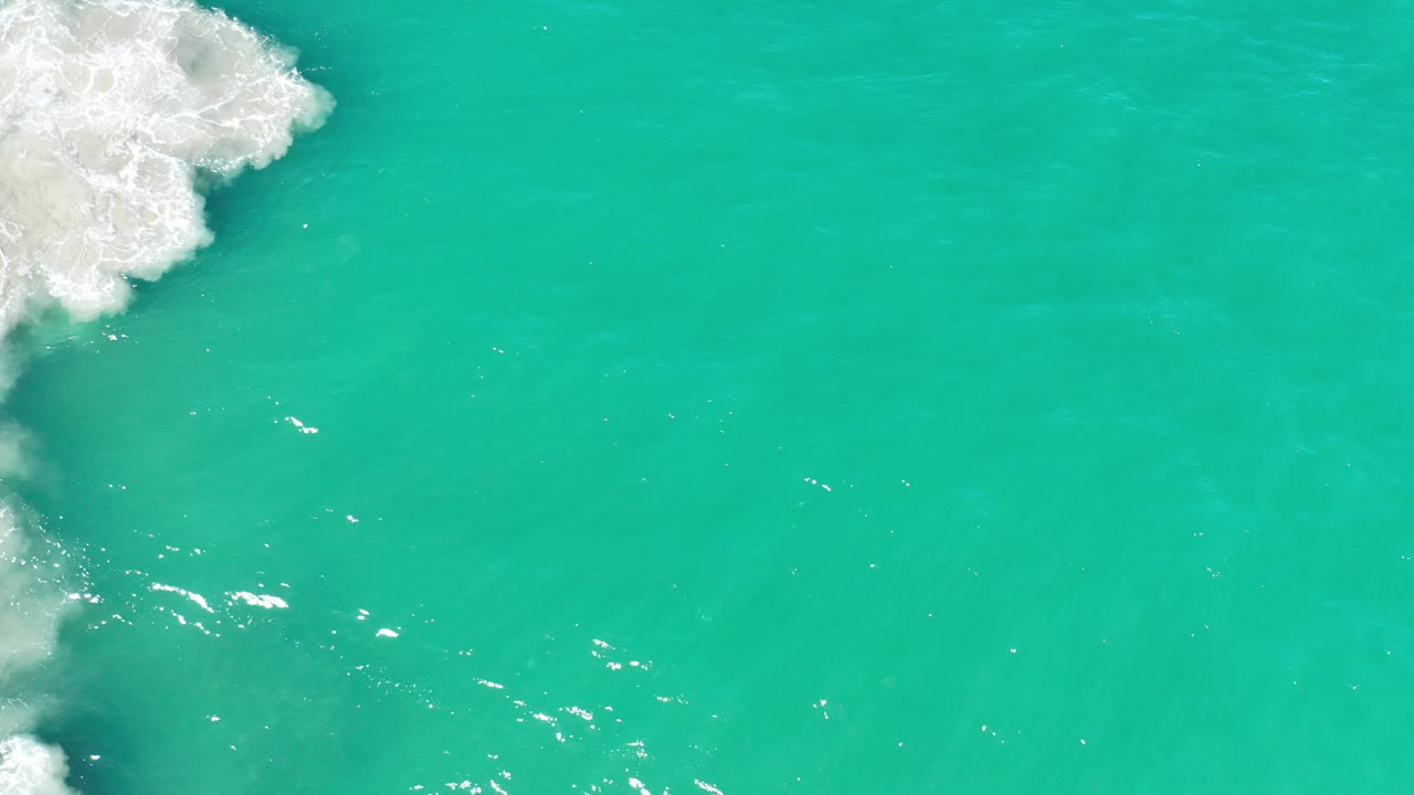 Overhead view of turquoise-green sea in Los Cabos with white surf clipped at the upper left edge, sun flecks on the water, and no visible people.