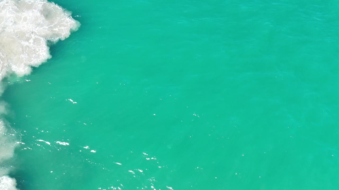 Overhead Los Cabos sea surface with a larger patch of white foam along the left edge, open green water across the frame, and no visible people.