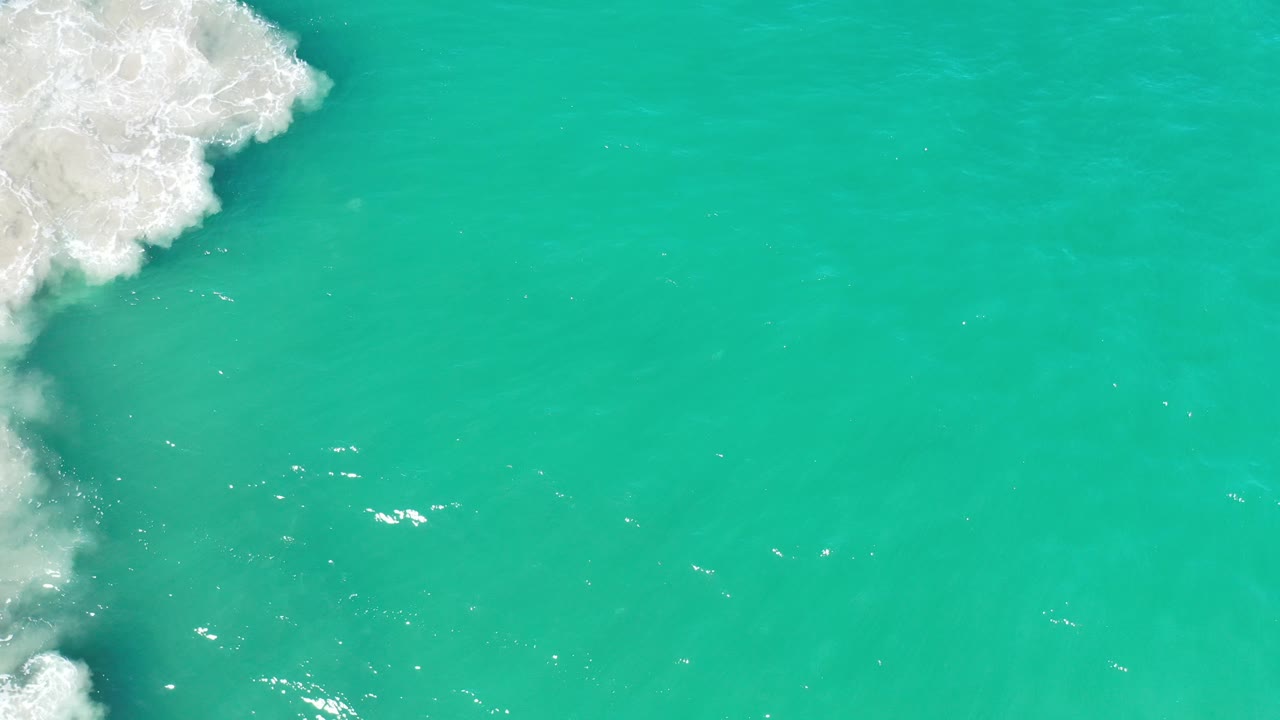 Aerial-style view of turquoise Los Cabos water with white surf concentrated in the upper left corner, scattered highlights, and no visible people.