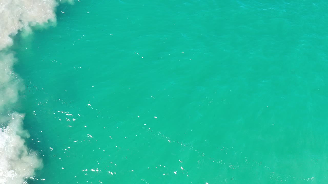 Overhead view of teal ocean water in Los Cabos, Mexico, with white foam entering from the upper left and no visible people.
