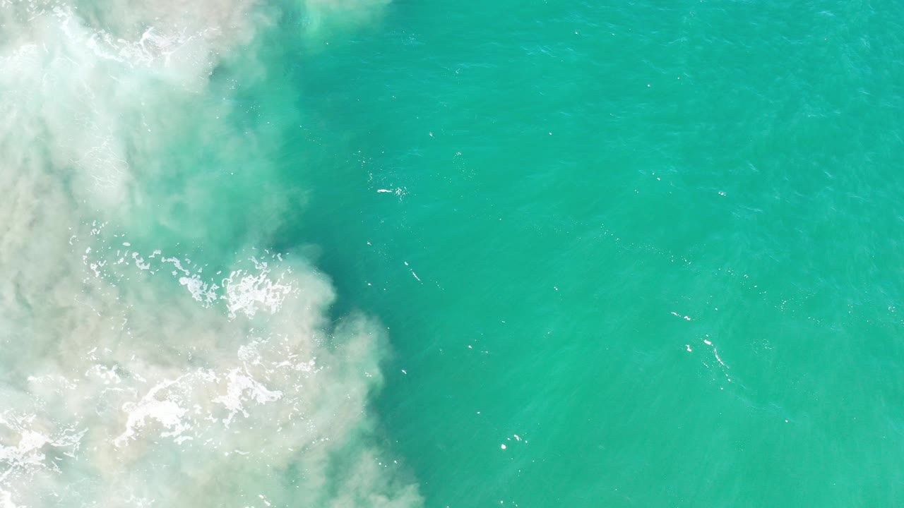 Overhead view of turquoise water in Los Cabos, Mexico, with white surf foam entering from the left side and no visible people.