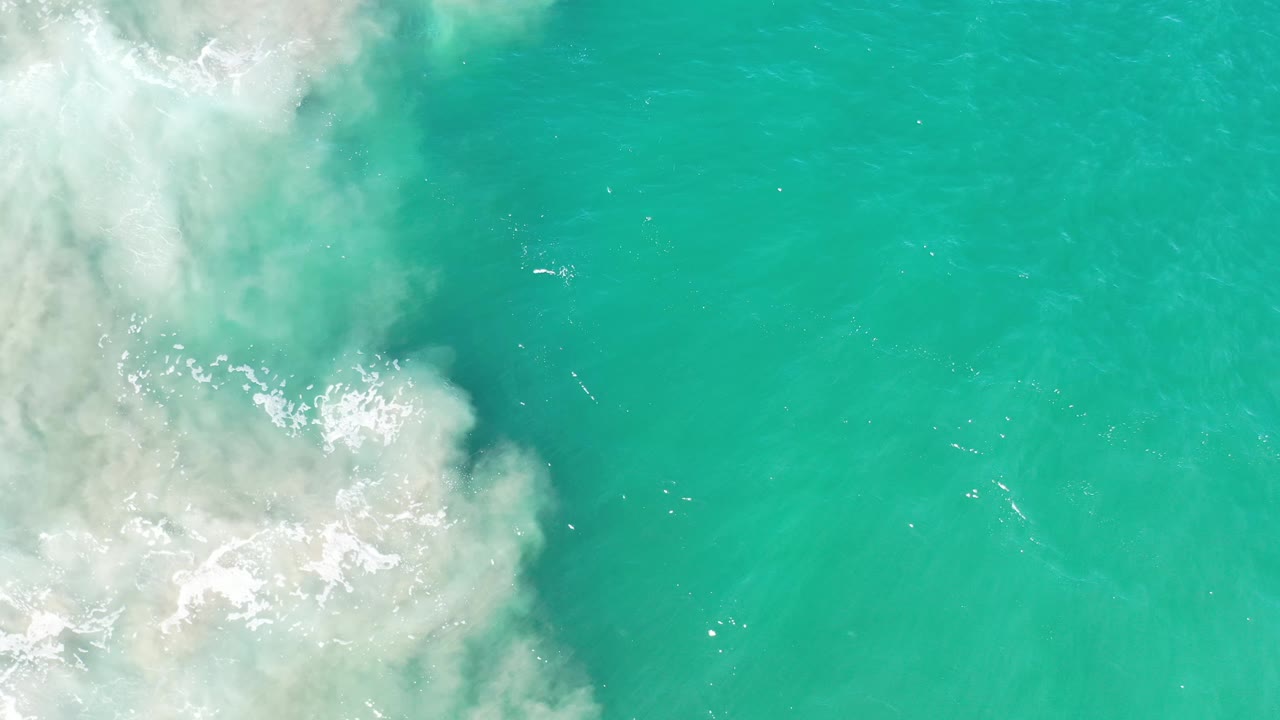 Overhead Los Cabos water frame showing a broader patch of white foam on the left and clear turquoise sea across the right, with no visible people.