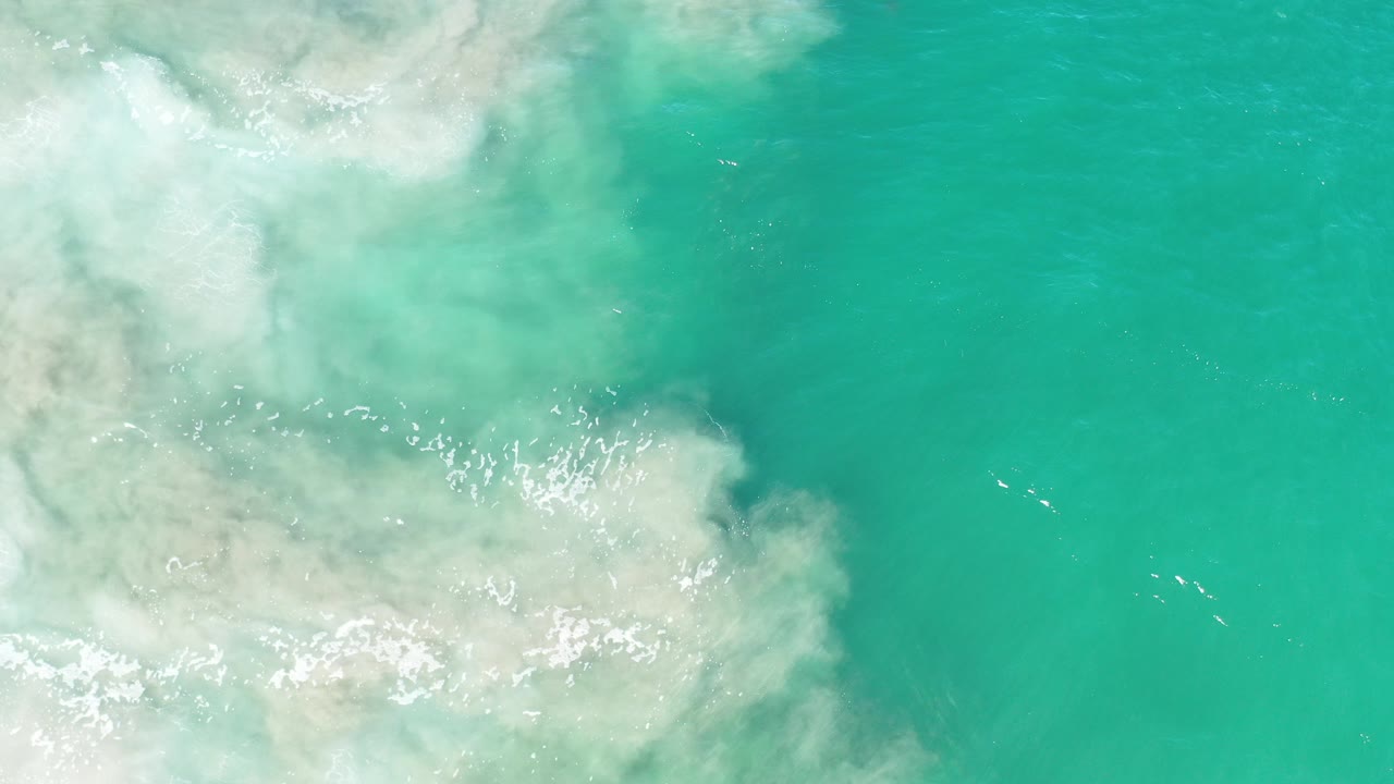 Overhead view of turquoise-green water in Los Cabos, Mexico with cloudy white foam spreading from the left edge and no visible people.