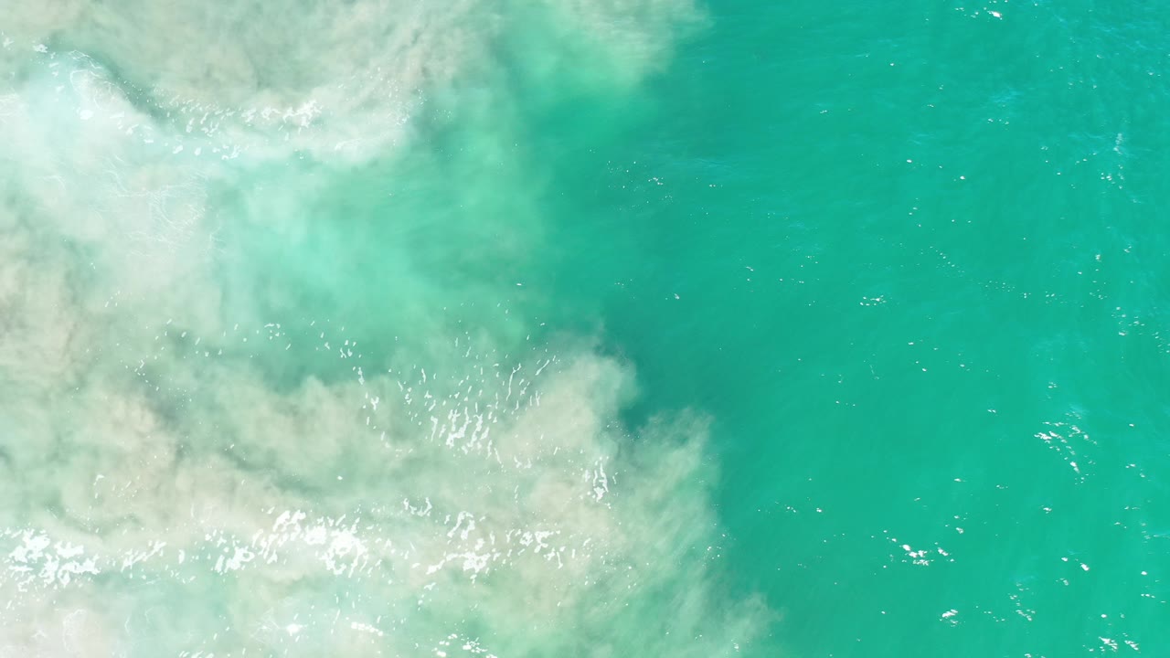 Overhead view of turquoise-green water in Los Cabos, Mexico, with pale wave foam and cloudy sediment on the left and no visible people.