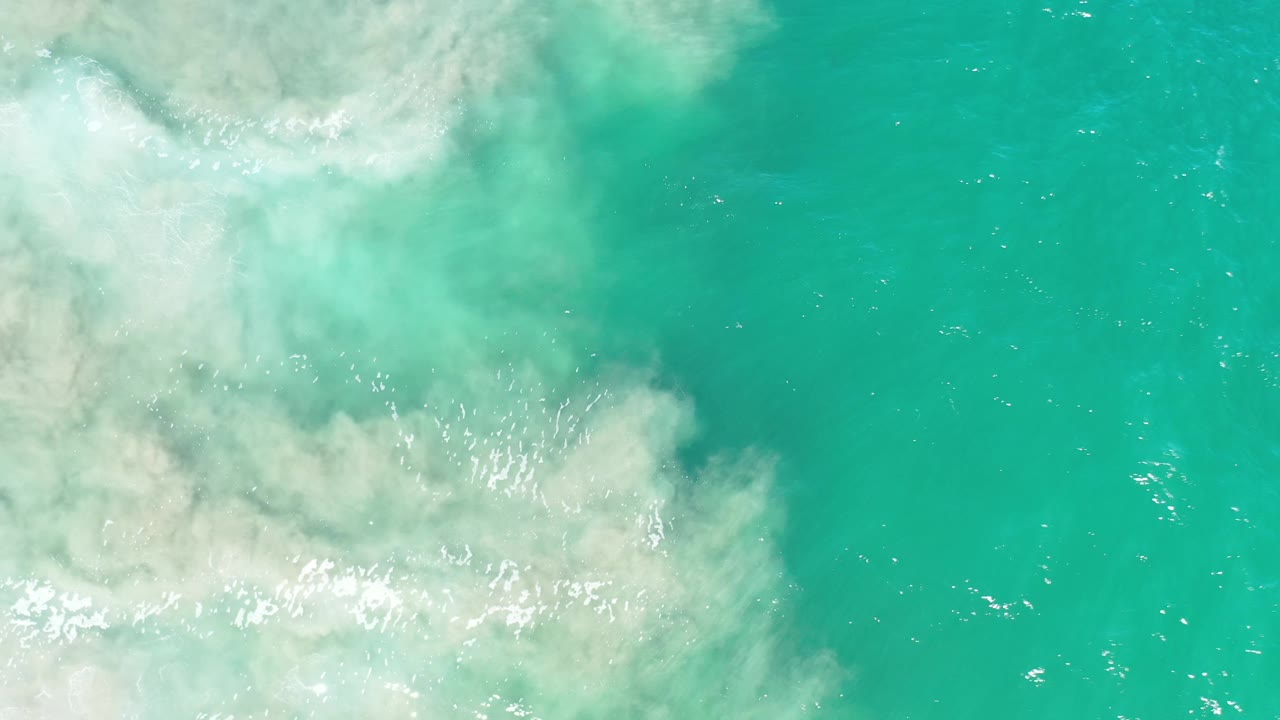 Overhead Los Cabos water view with a broad pale foam patch at upper left, clear green water to the right, and no visible people.