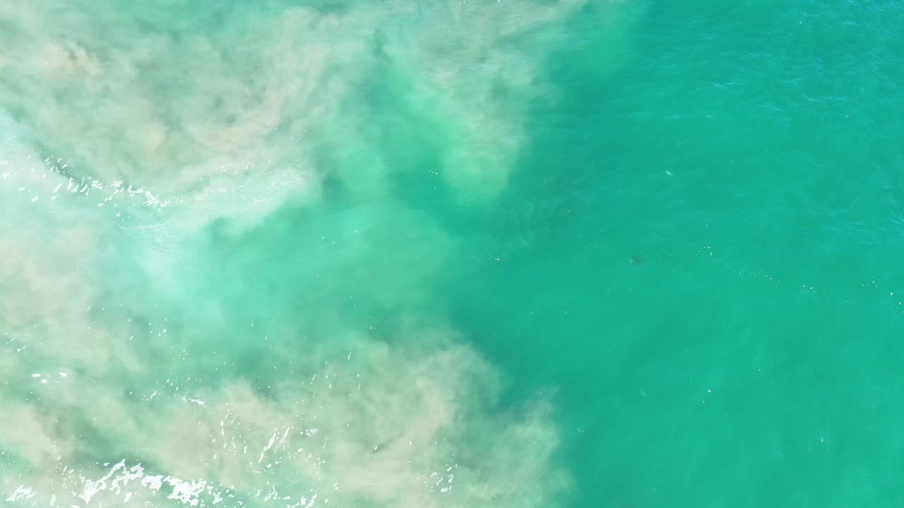 Overhead view of turquoise Los Cabos water with pale suspended sand spreading from the left side and no visible people.