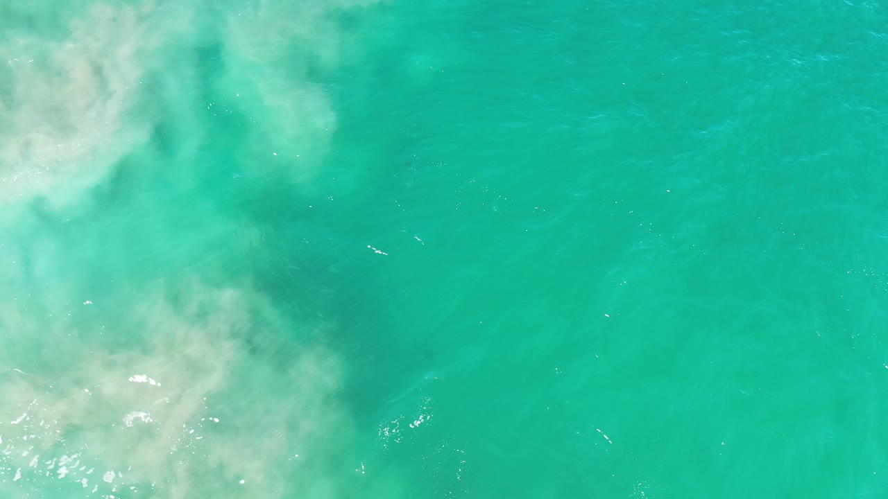 Overhead view of turquoise ocean water in Los Cabos, Mexico, with a broad white foam patch entering from the upper left and no visible people.